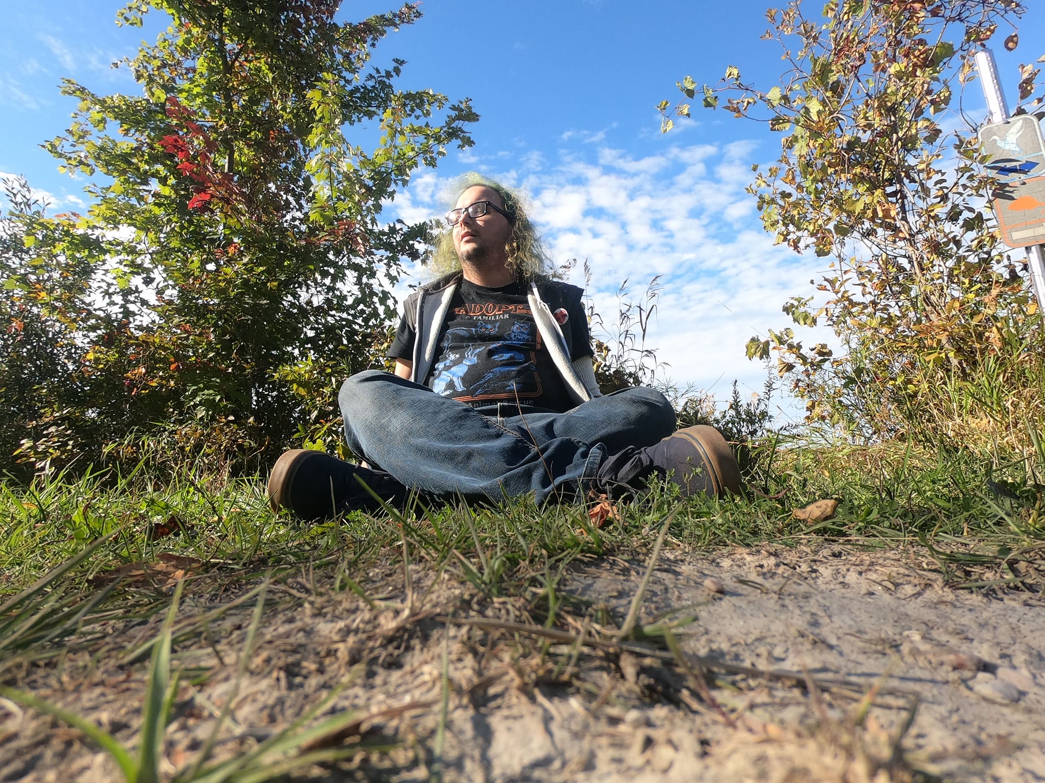 A man with long green hair sits on the ground, looking tired, on the side of a path through a wetland